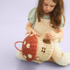A young child examines a Olli Ella Red Mushroom Basket, woven in red and white, sitting on a light purple background.