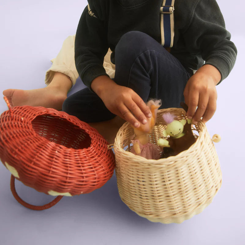 A child sits on the floor, playing with toys in a Olli Ella Red Mushroom Basket, alongside another empty decorative basket. The child is wearing a green hoodie and light pants. The background is plain and light.
