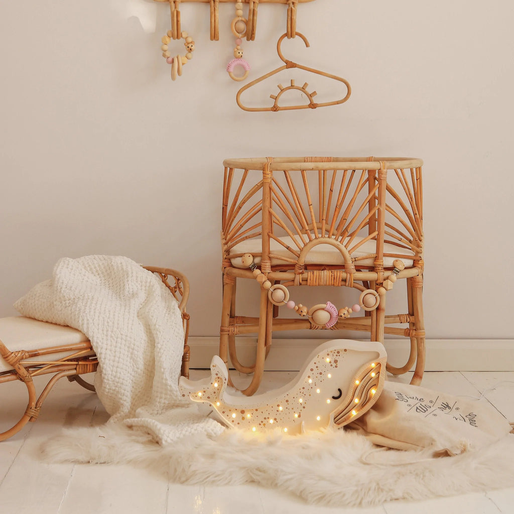 A cozy nursery corner featuring a wooden rattan chair and stool, a soft white rug, a glowing Sharky Lights Whale Lamp, and hanging wooden baby toys and hangers.