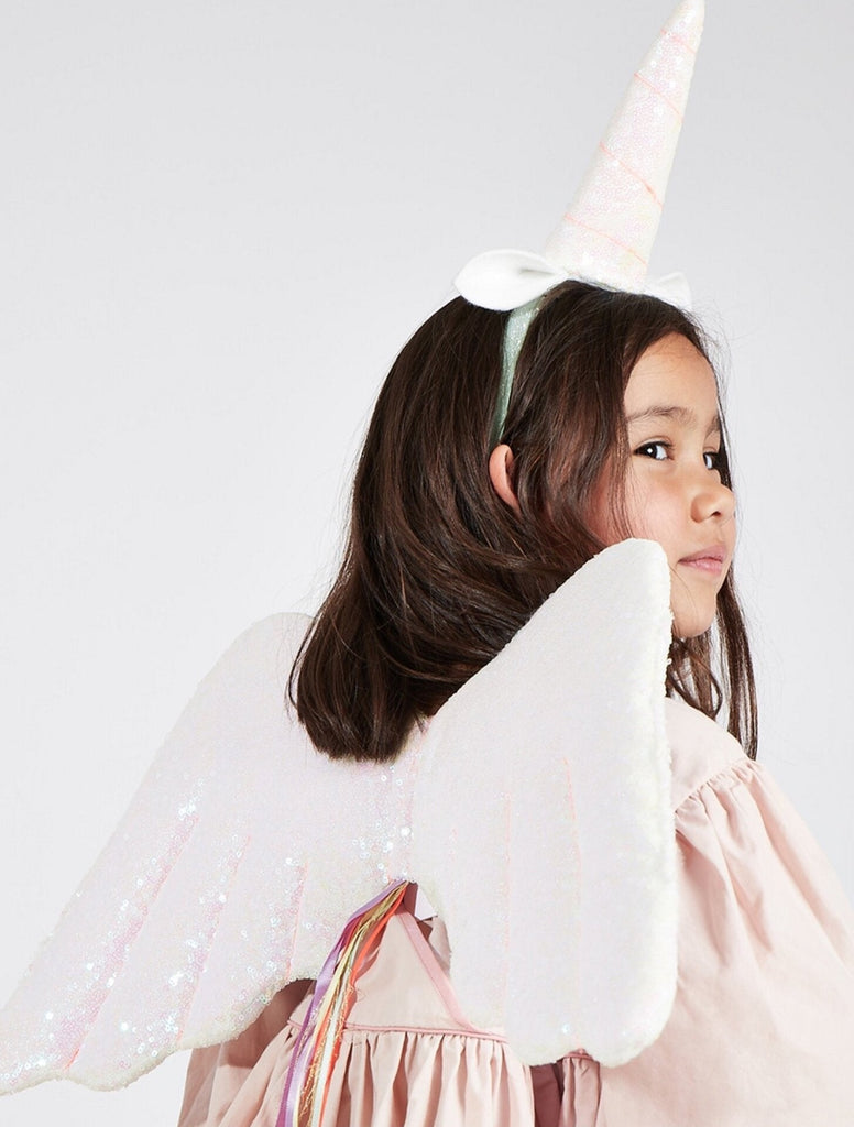 A young girl wearing a Meri Meri Winged Unicorn Costume with a horn headband and shimmering wings, looking to the side against a light background.