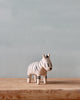 A Tiny Wooden Zebra figurine stands on a wooden surface against a plain, light blue background. The zebra is painted with black stripes and has a simplistic, stylized design.