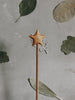 A Handmade Wooden Magic Wand, possibly made of cherry wood, topped with a star-shaped ornament against a light gray background. There are several dried leaves of different shapes and shades of green scattered around the background, creating a natural and rustic setting.