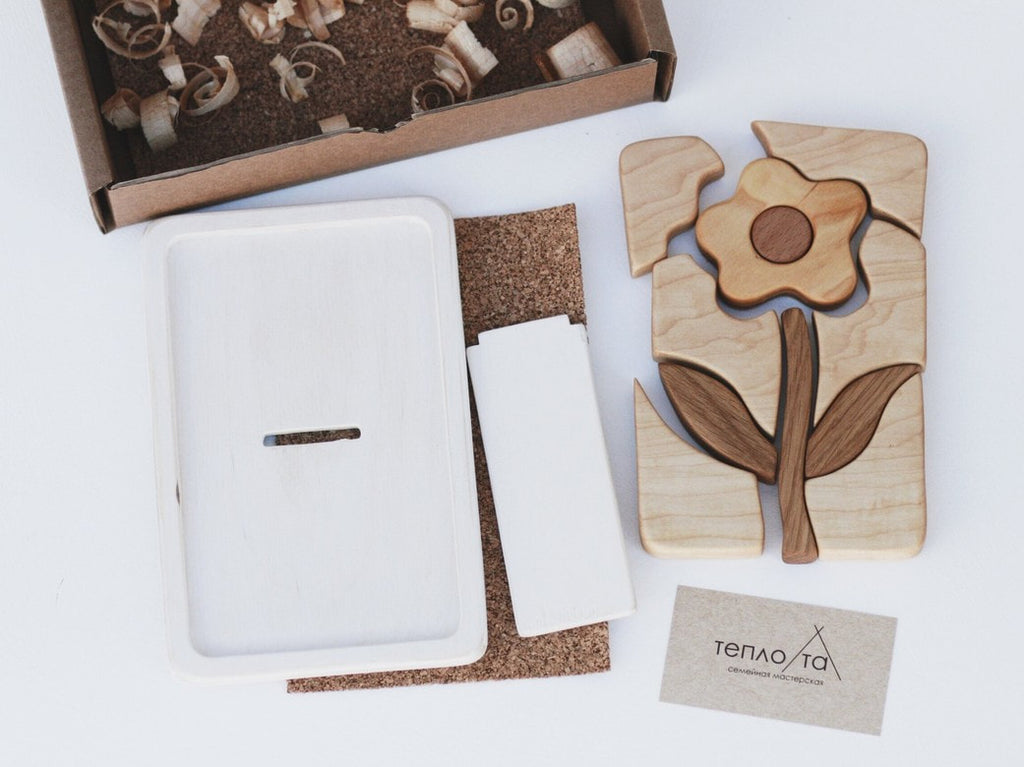 A Handmade Wooden Puzzle, handmade in Russia from Cherry wood and finished with linseed oil, depicts a flower with leaves, disassembled on a white surface. Nearby is a white rectangular stand with a slot, a piece of cork material, a card with text, and a box containing wood shavings.