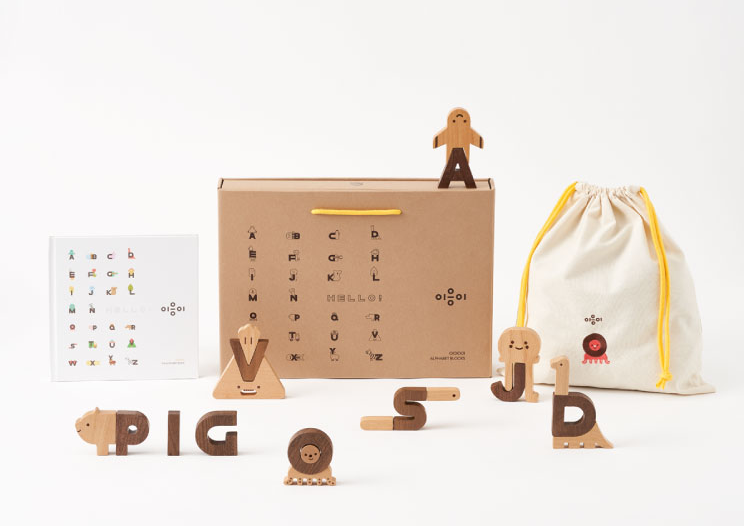A children's Ultimate Wooden Alphabet Puzzle displayed against a white background. It includes a wooden alphabet set, a box with letter cutouts, and a cloth bag. Some letters are arranged to spell "pig" and