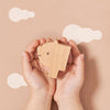 Two hands holding a Ultimate Wooden Alphabet Puzzle with a simple face drawn on it against a beige background with white paper clouds and wooden alphabet blocks.