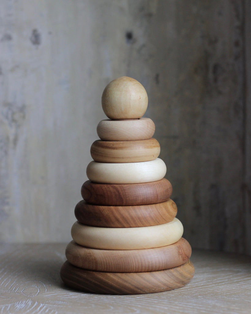 A wooden stacking toy with wooden rings in various natural shade of wood, stacked from large to small, topped with a sphere shaped wooden piece.