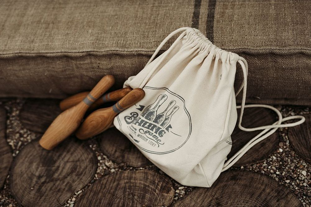 Beige canvas bag with strings to close it. 3 wooden bowling pins laying next to it. 