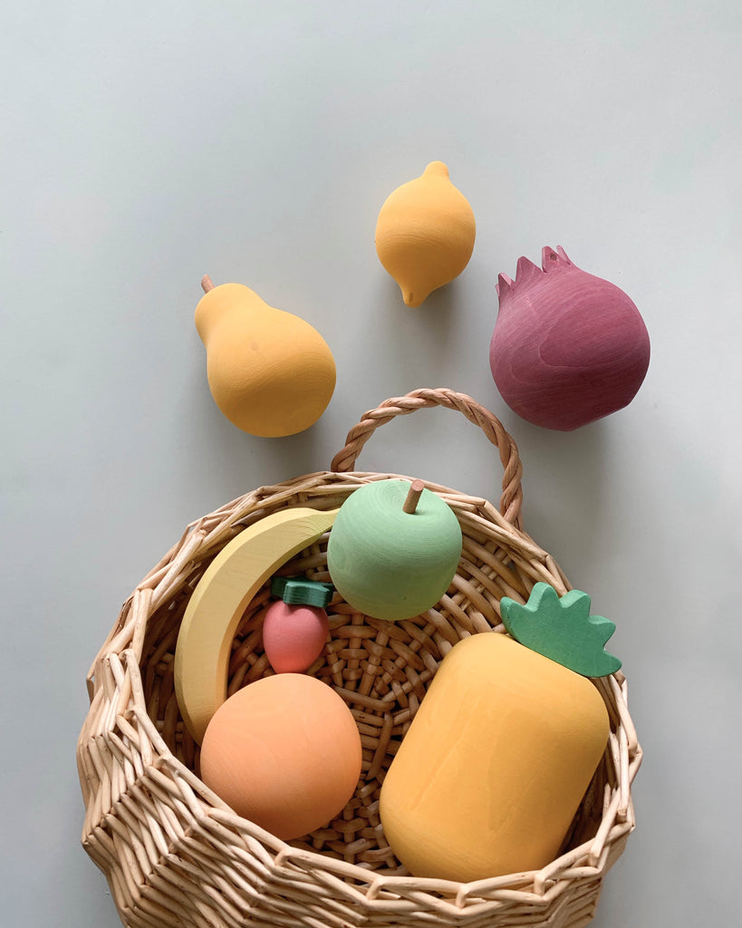 A wicker basket filled with Raduga Grez Handmade Painted Wooden Fruits including a yellow lemon, green apple, pink strawberry, orange peach, and a few others on a gray background.