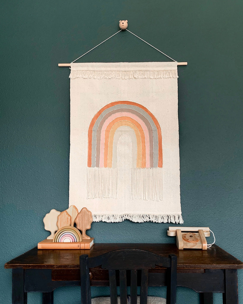 A Handmade Rainbow Wall Hanging with a rainbow design in earth tones on a dark green wall, above a small wooden table with decorative items that match the artwork's theme.