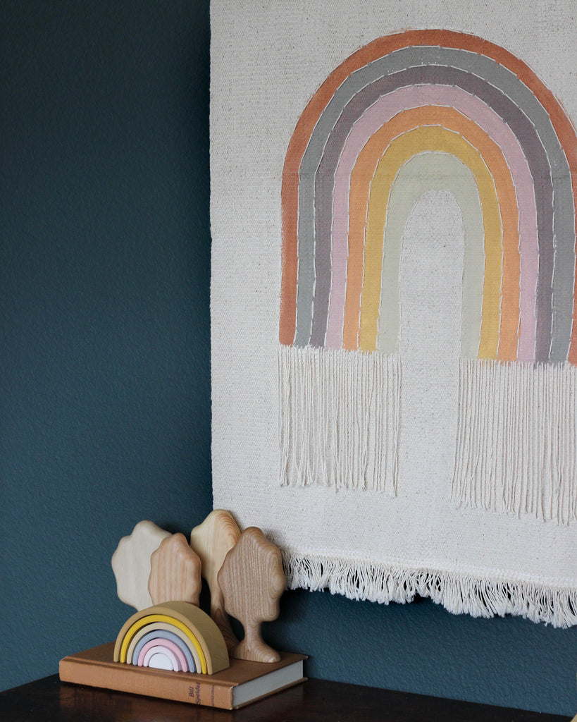 A Handmade Rainbow Wall Hanging hangs on a dark green wall next to a wooden bookend shaped like clouds, supporting a stack of books.