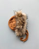 A hand-knit The Blueberry Hill Lion Hat with a faux fur trim and braided ties, placed on a plain light grey background.