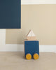 A Sea & Boat Push Toy hand-made wooden toy car painted blue with yellow wheels and a small triangular block placed on top, set against a backdrop of a beige and navy geometric canvas.