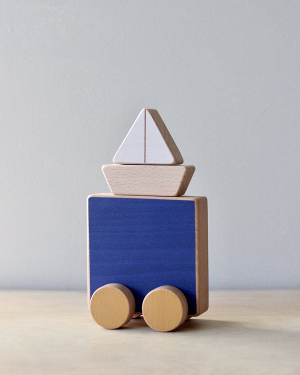 A simple eco-friendly Sea & Boat Push Toy, crafted from FSC certified beech wood, with a blue body and natural wood wheels, topped with a stack of geometric wooden blocks shaped like a sailboat
