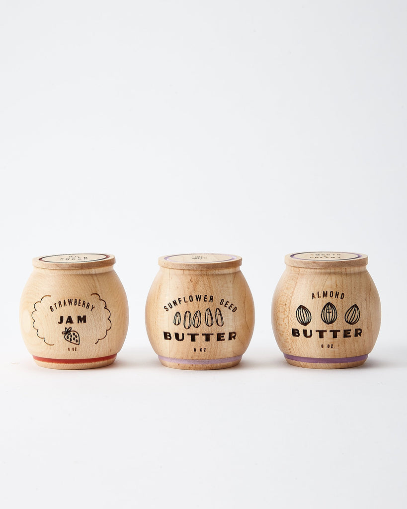 Three wooden jars labeled "Milton & Goose Jam & Nut Butter Pretend Play Food," "Sunflower Seed Butter," and "Almond Butter" in decorative script, lined up against a white background.