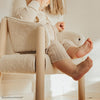 A young child sitting in a cozy, white, Charlie Crane SABA Chair 'Fur' made from raw beech wood, dressed in a beige knit outfit. The child's face is not visible, and one hand is up touching their mouth, with their legs relaxed and feet visible in the warm, softly lit room.