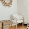 A cozy, small white Charlie Crane SABA Chair 'Fur' with a raw beech wood frame sits in the corner of a room. A wicker basket and a woven mat are on the wooden floor nearby, and a round mirror with a decorative rattan frame hangs on the wall above the chair.