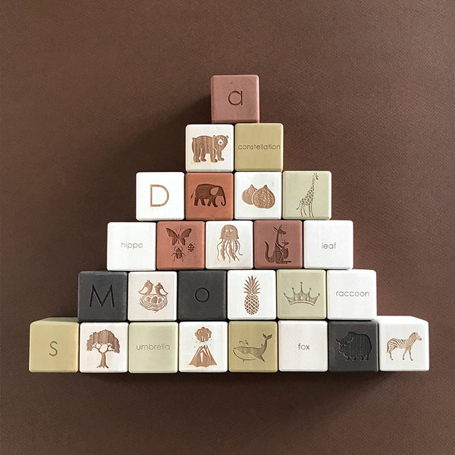 A pyramid of Alphabet Wooden Blocks - Olive, each crafted from natural linden wood and illustrated with a letter and corresponding animal or object, placed on a brown background.