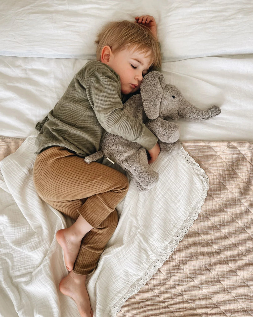 A young child asleep on a bed, hugging a Senger Naturwelt Cuddly Animal - Elephant plush toy. The child is wearing a green shirt and brown pants. The setting is cozy.