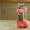 A small hand-felted grey mouse holding a pink heart in its arms. Photographed sitting on a peach.