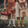 Two children in vintage clothing, one holding an Olli Ella Red Mushroom Basket and the other a picnic basket, standing in a forest with flora details.