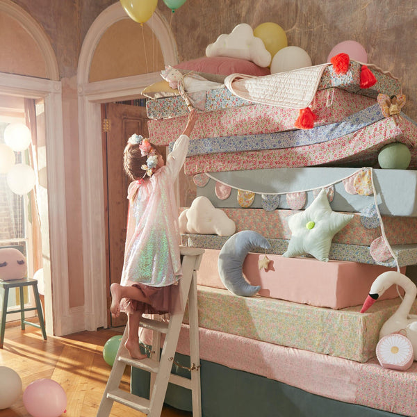 A young girl in a sparkly dress with the Meri Meri Iridescent Sequin Cape Costume reaches up to bunk beds adorned with colorful bedding and plush toys, surrounded by balloons, in a warmly lit room.