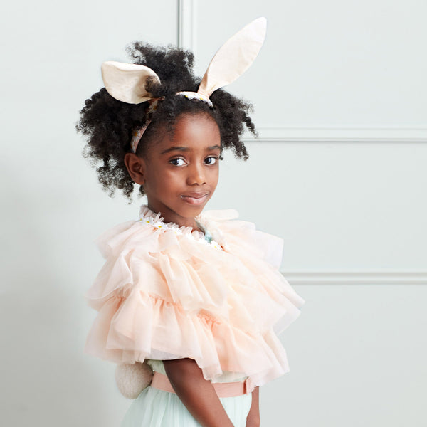 A young girl with curly hair wearing a Meri Meri Peach Tulle Bunny Costume, looking towards the camera with a gentle smile, standing against a light grey background.