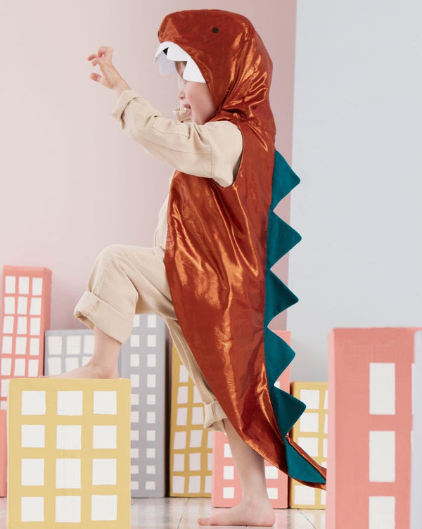 A child in a Meri Meri Dinosaur Costume playfully poses among colorful cardboard buildings. The costume is brown with a teal spine.