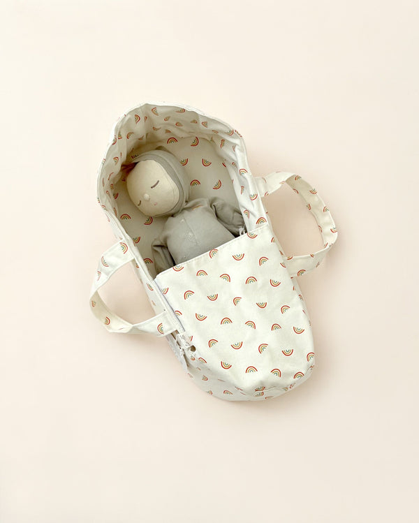 A beige Olli Ella Rainbow Carry Cot with a toy inside, featuring a rainbow patterned interior, photographed on a light beige background.