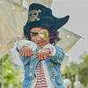 A child dressed in a Meri Meri Pirate Costume with a large hat, eye patch, and denim jacket, extends fists with "ahoy mate" written on them, smiling playfully outdoors.