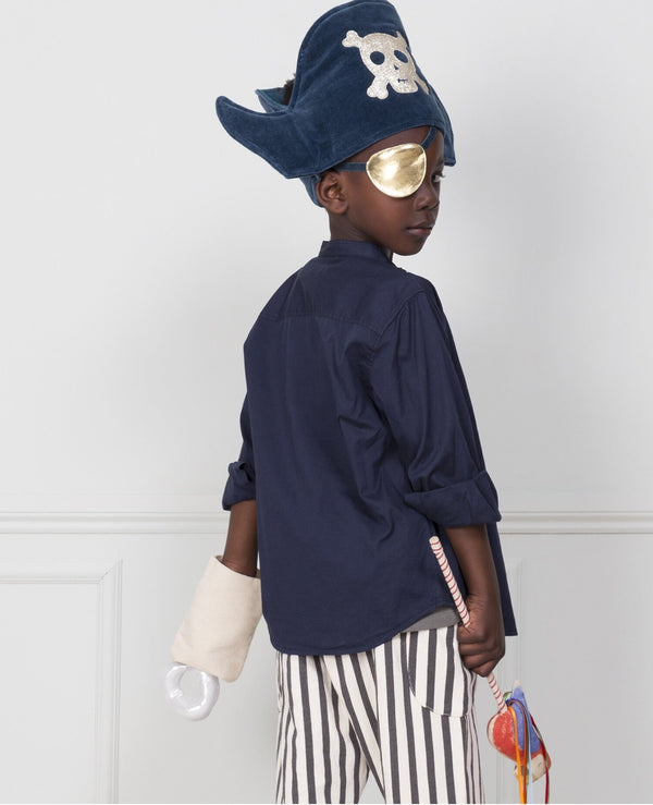 An imaginative play unfolds as a young boy in a Meri Meri Pirate Costume - Final Sale, featuring a blue hat with a skull, eye patch, striped pants, and a hook, stands against a white wall, looking over his
